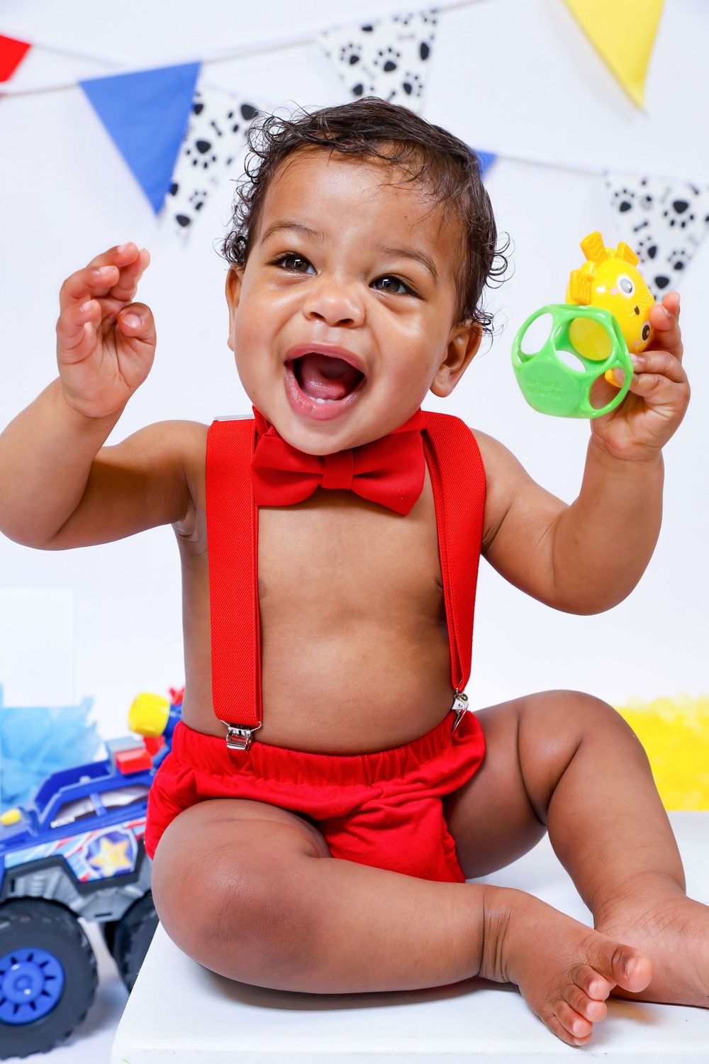 Baby wearing red outfit sits joyfully with colorful party decorations in background.