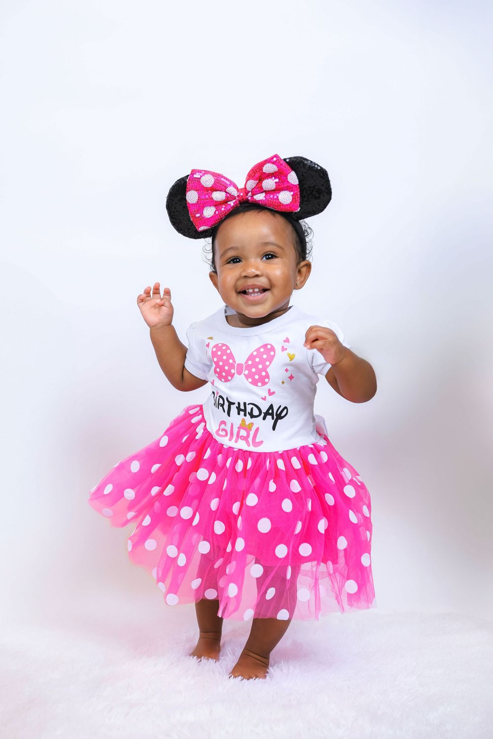 A toddler dances in a pink polka dot tutu dress and Minnie Mouse ears against white background.