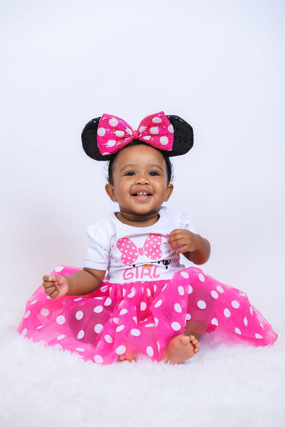 A happy toddler wearing a pink polka dot Minnie Mouse costume sits on a white backdrop.