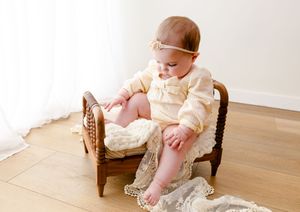 Baby wearing cream sweater sits in small vintage wicker chair on hardwood floor with lace fabric.