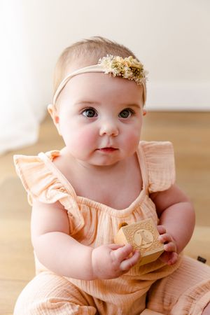 Sweet baby in peach-colored ruffled dress with gold headband smiles while sitting on wooden floor.