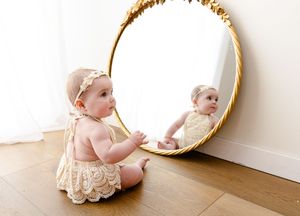 A baby in a cream lace dress crawls near a circular mirror on wooden flooring, shown in multiple sequential shots.