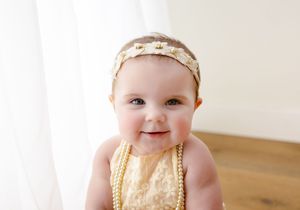 Baby wearing a gold sequined dress and white headband sits on wooden floor during photo session.