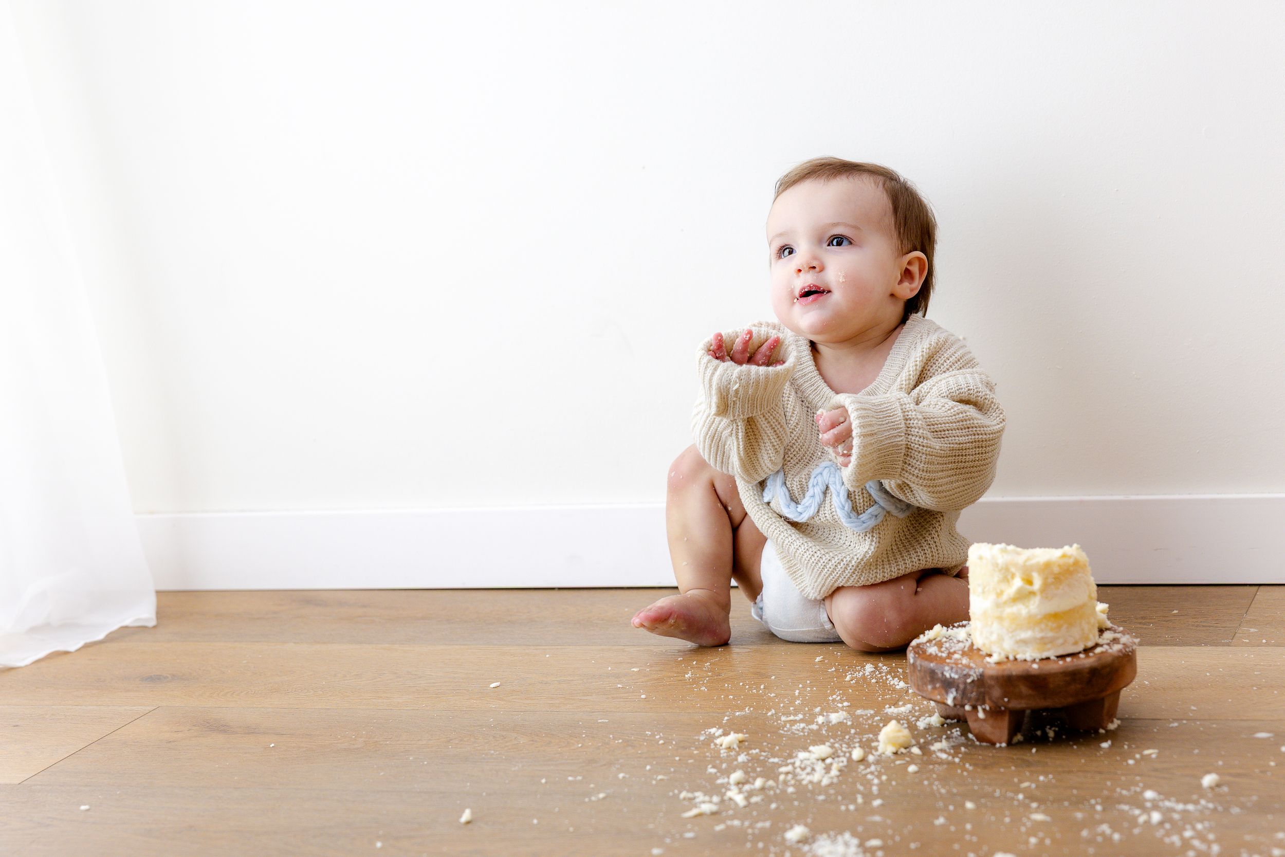 A baby in a white outfit sits at a table with a birthday cake and smashed frosting mess.