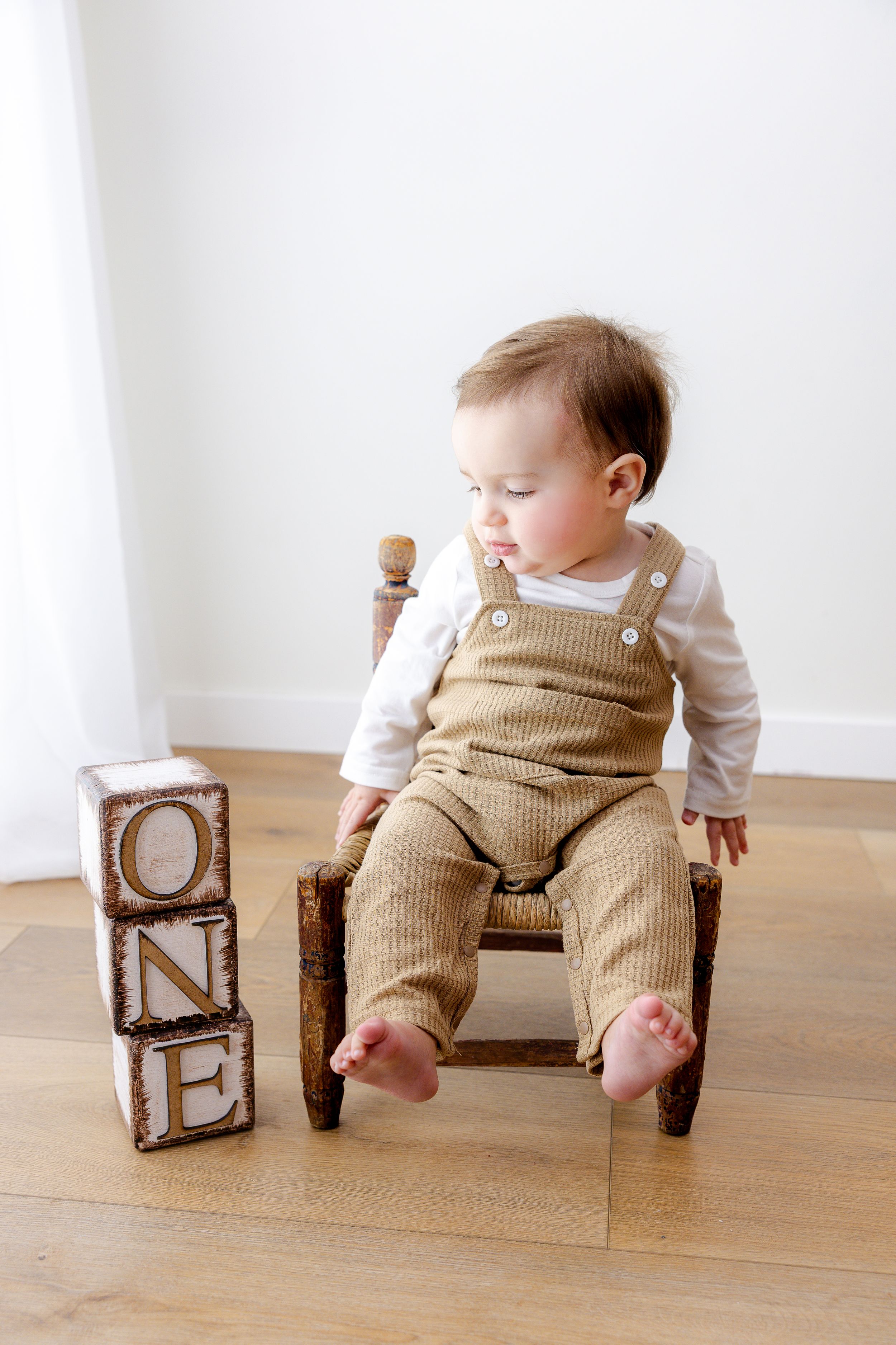 Baby wearing tan overalls sits between wooden alphabet blocks on hardwood floor.