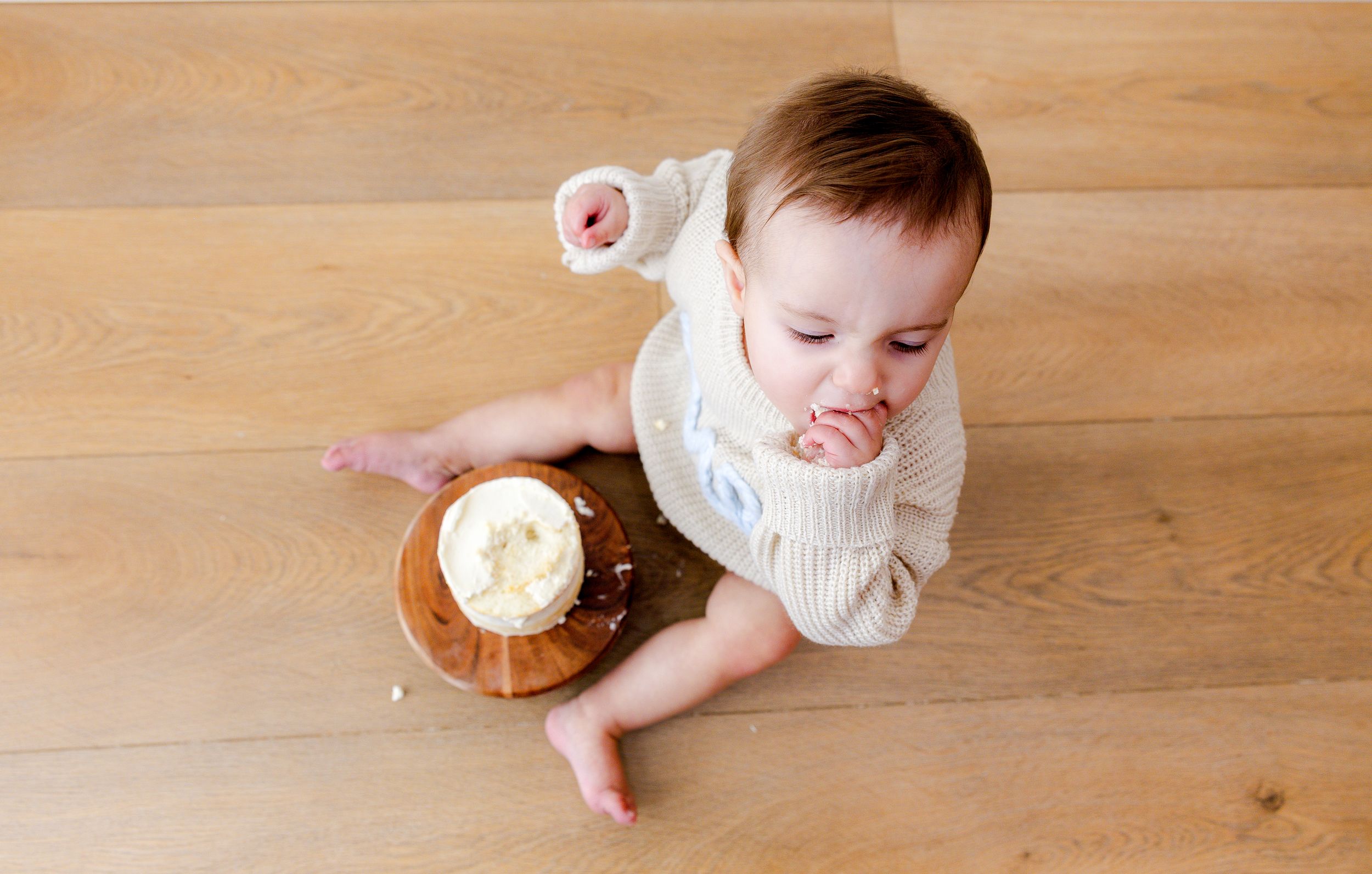 Baby in white sweater sitting on wooden floor with smashed cake during first birthday cake smash photo session.