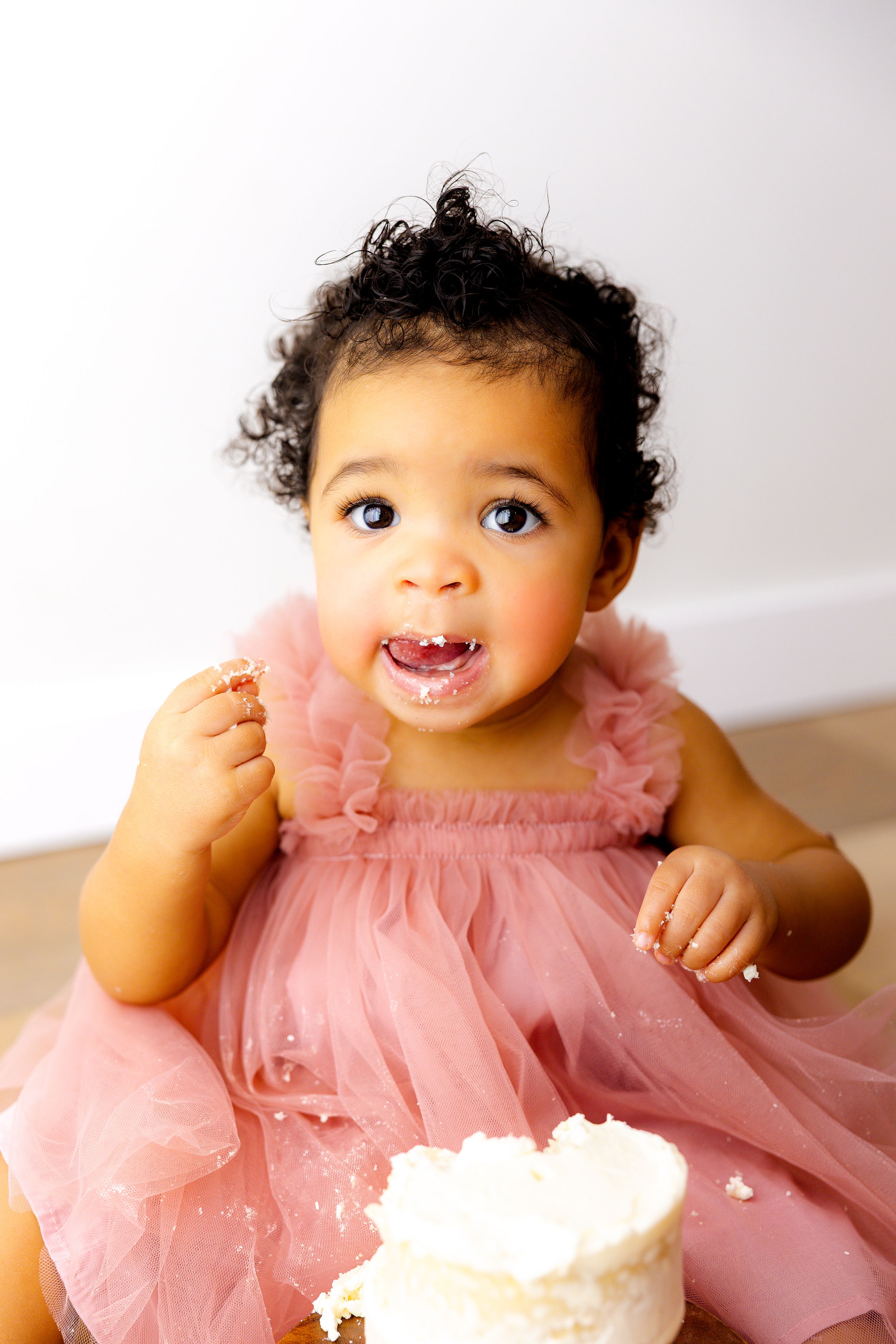 Baby in pink tulle dress smiles while enjoying birthday cake smash photo session.