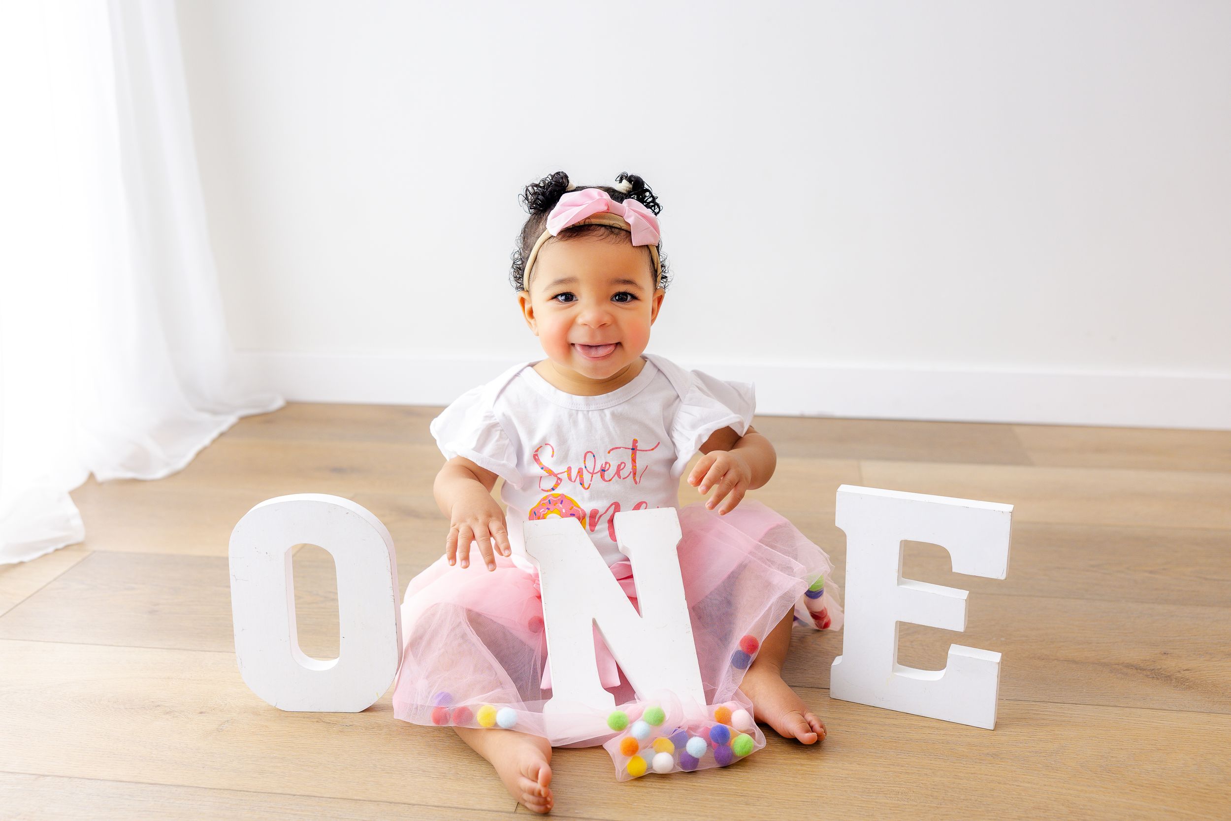 Baby in pink tutu dress sits on wooden floor with decorative white letter blocks spelling out 'ONE'.