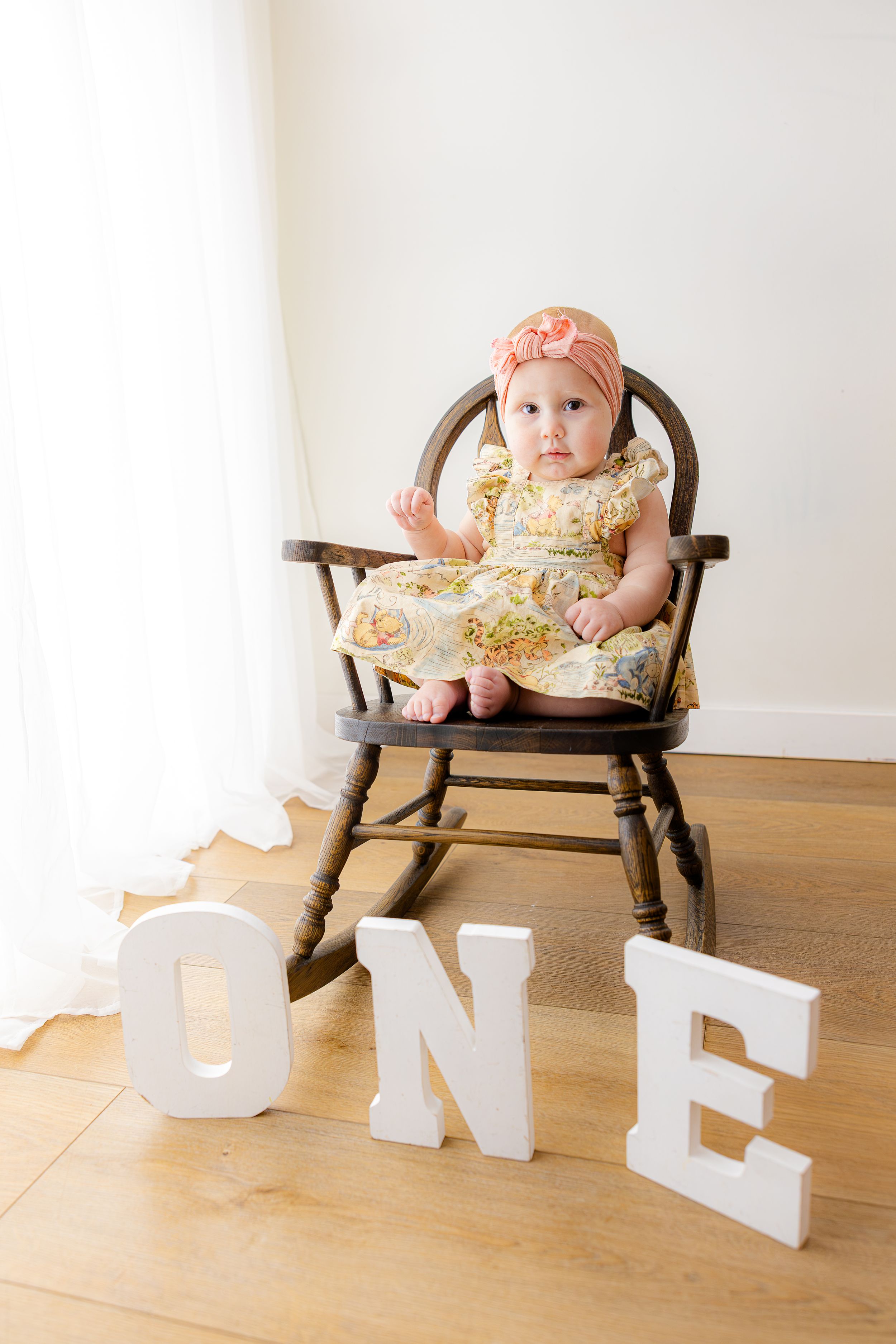 Baby in a wooden chair with decorative white letters spelling 'ONE' arranged on hardwood floor.
