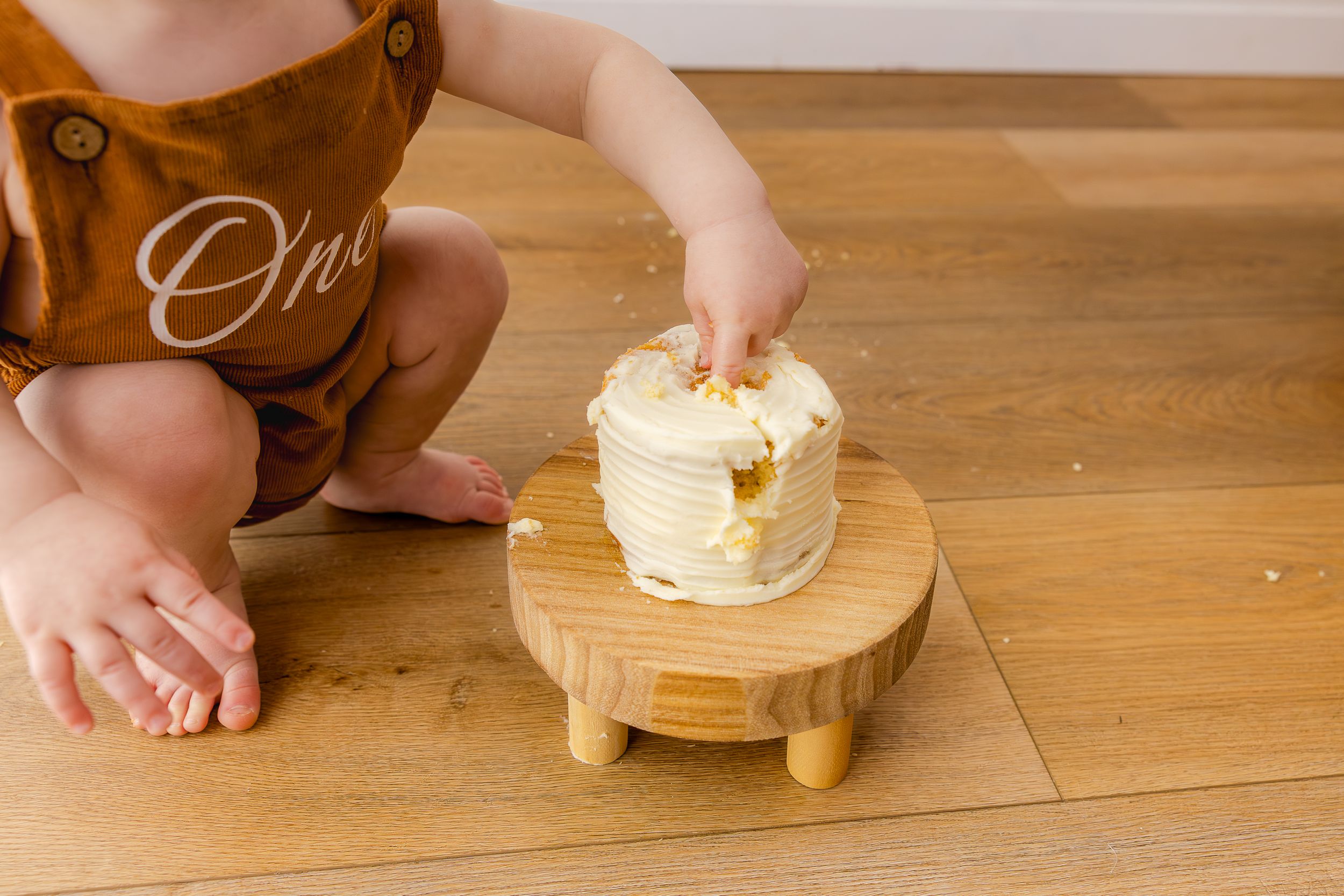 Baby in 'One' outfit smashing a white frosted smash cake on a wooden stand during first birthday celebration.