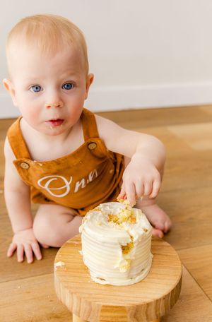Blue-eyed baby in brown romper smashing a white frosted smash cake on a wooden board during first birthday.