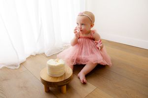 Baby in pink tulle dress sits on hardwood floor next to small wooden cake stand with white birthday cake.