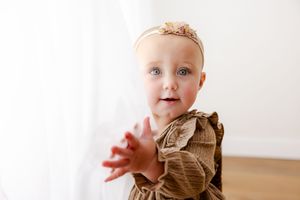 Baby in brown dress reaches forward while sitting on light wooden floor during portrait session.