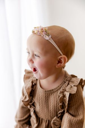 Baby in brown ruffled sweater and delicate headband smiles against white background.