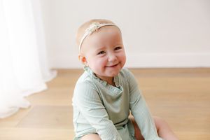 Baby in mint green sweater sits on wooden floor with bright smile and happy expression.