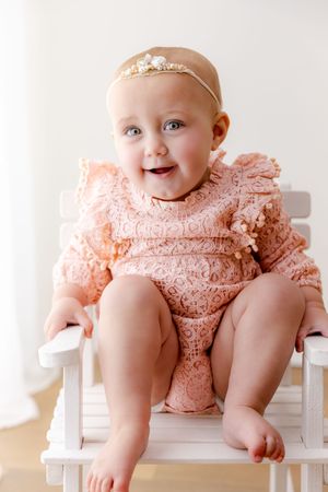 Baby wearing peach lace dress sits on white wooden bench during portrait session.