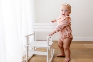 A baby in a peach-colored romper stands near a white wooden chair on hardwood flooring.