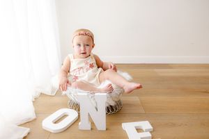 Baby in white onesie sits on wooden floor with decorative letters spelling 'ONE' for first birthday milestone photo.