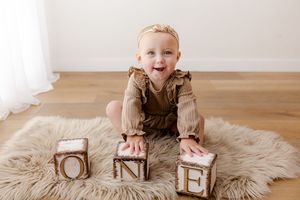 Baby wearing brown sweater lies on fluffy rug next to wooden alphabet blocks showing joyful expression.