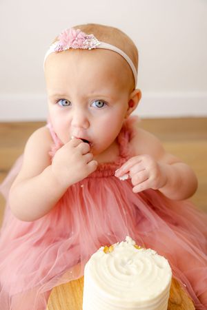 Baby in pink tulle dress reaches towards white frosted cake during first birthday photoshoot.