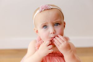 Small child in pink dress and headband with hands near mouth against white wall.