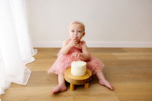Small child in pink dress sitting on wooden floor next to white cake on pedestal.