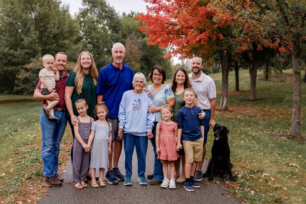 A group stands together outdoors in autumn with vibrant red maple trees and fallen leaves in the background.