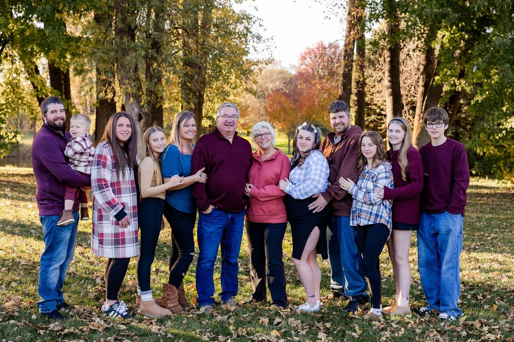 A group of people in casual attire stand together outdoors during autumn with colorful fall foliage in the background.