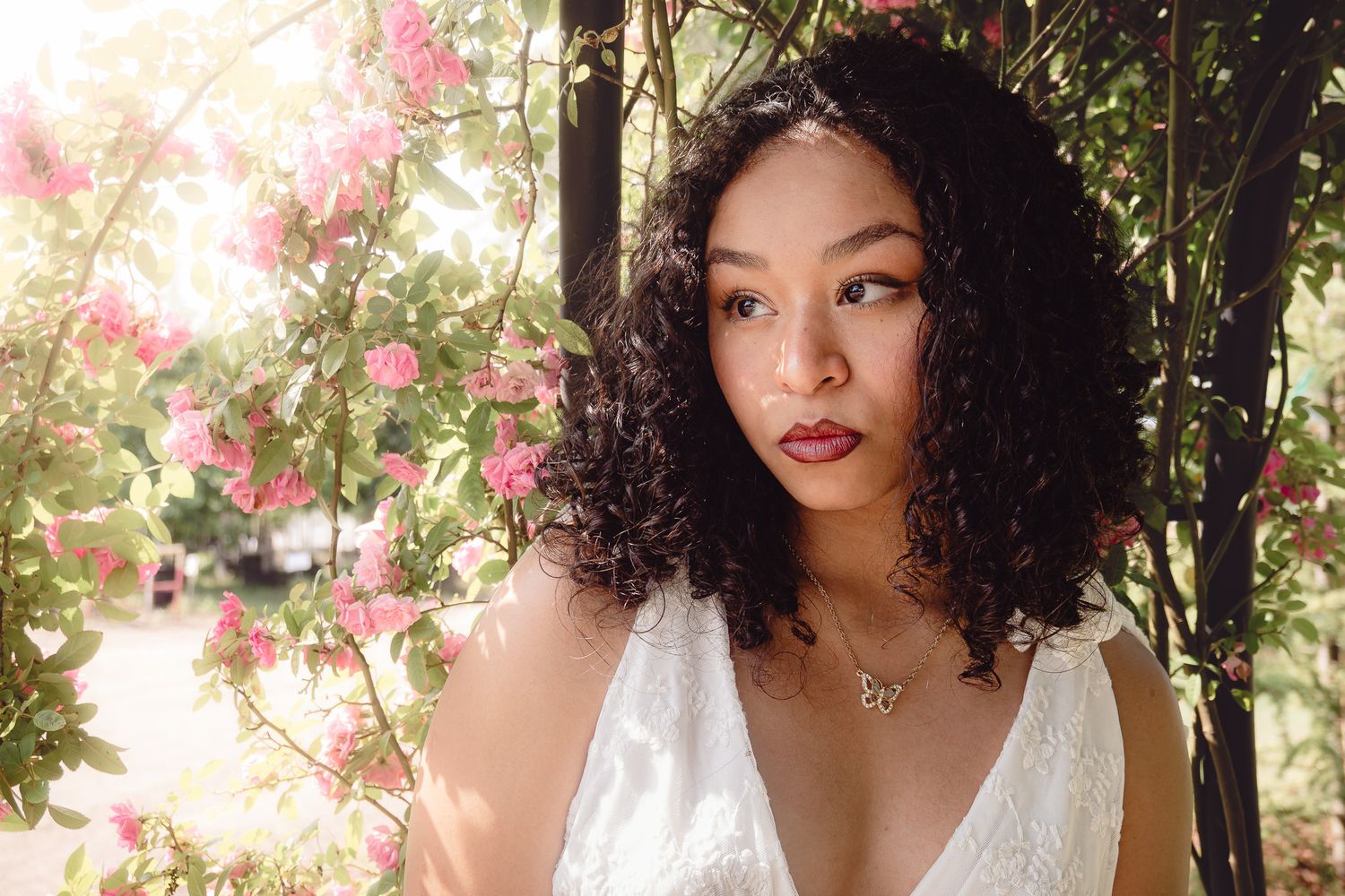 A person in a white dress poses outdoors among pink flowers and trees with natural sunlight creating a dreamy effect.