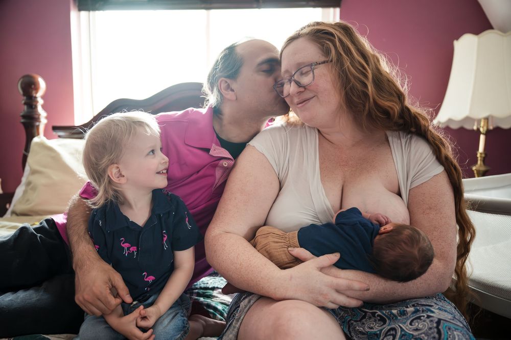 Family bonding moment with father kissing mother while she breastfeeds newborn, young blonde child sitting nearby.