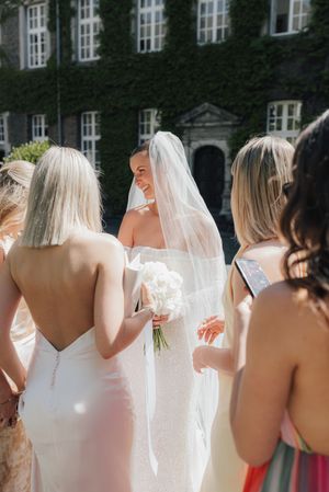 A wedding party stands outside an ivy-covered building with white windows on a sunny afternoon.