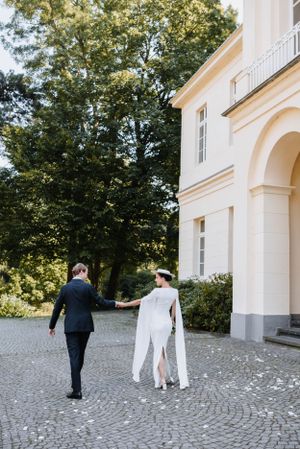 Newlyweds walk hand in hand on cobblestone path near white colonial building.