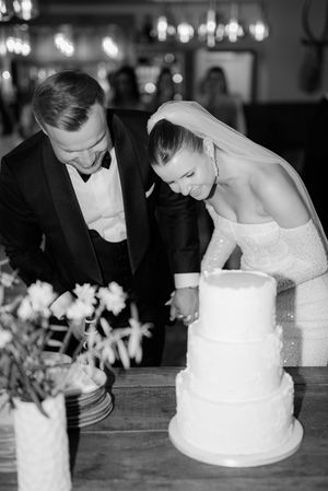 Couple in formal wedding attire looking down at a simple white three-tiered wedding cake during reception.