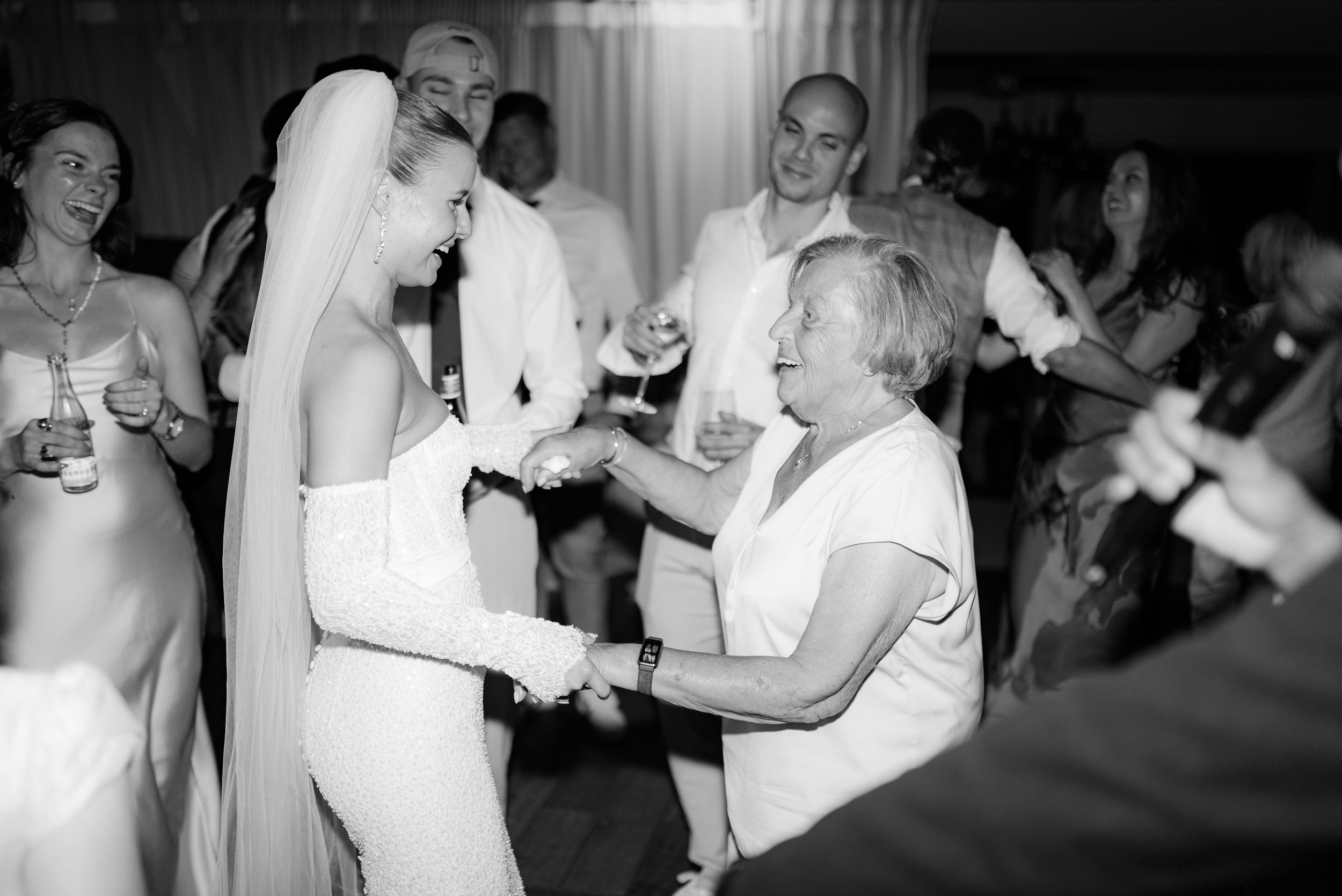 Black and white photo of joyful dancing at a wedding reception with a bride in a white dress and party guests.