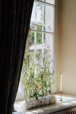 Delicate white flowers arranged in a windowsill marble planter with a black curtain and candlestick nearby.