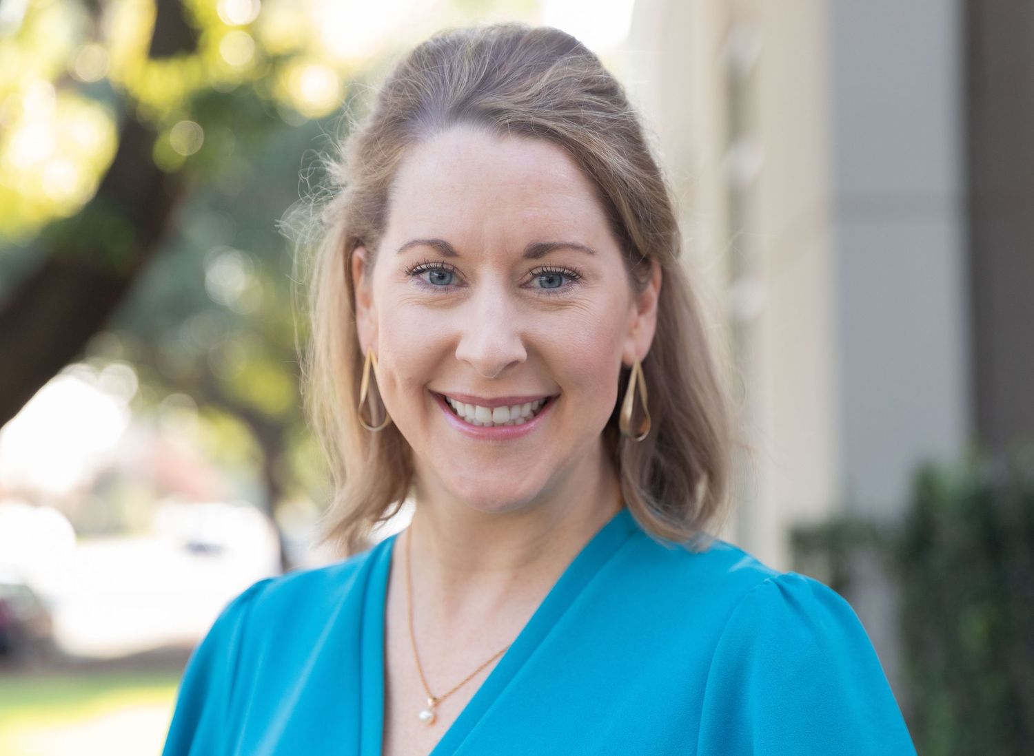 Professional headshot of a person in a bright turquoise blouse smiling warmly at the camera outdoors.