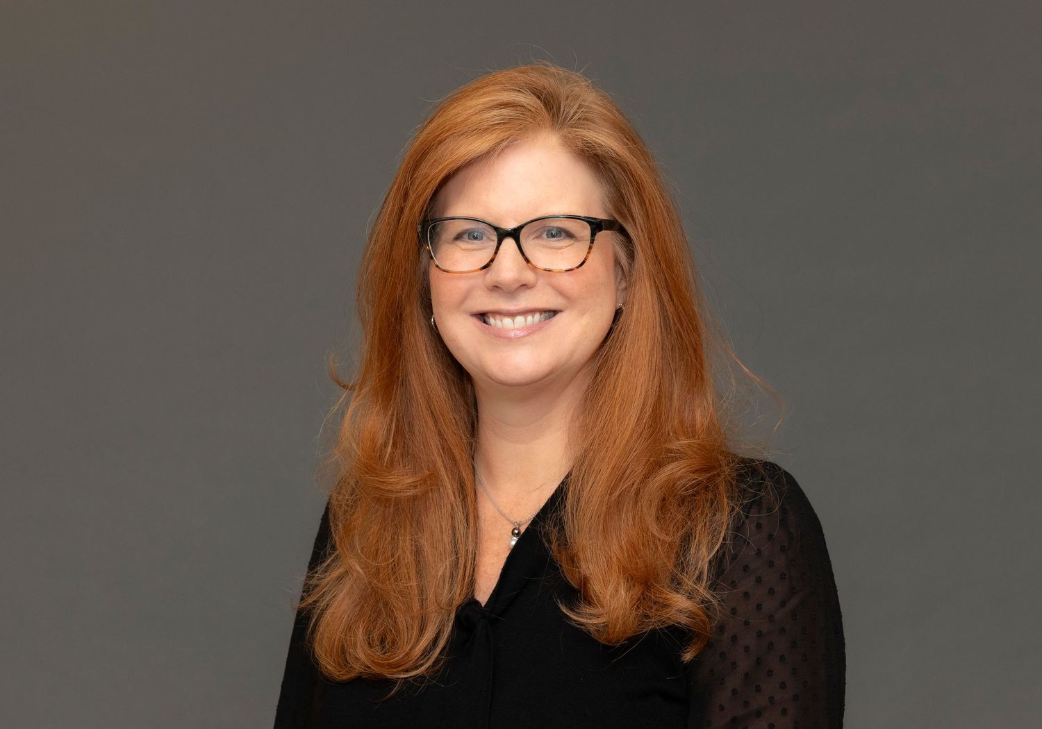 Professional headshot of a person with long red hair wearing glasses against a grey background.