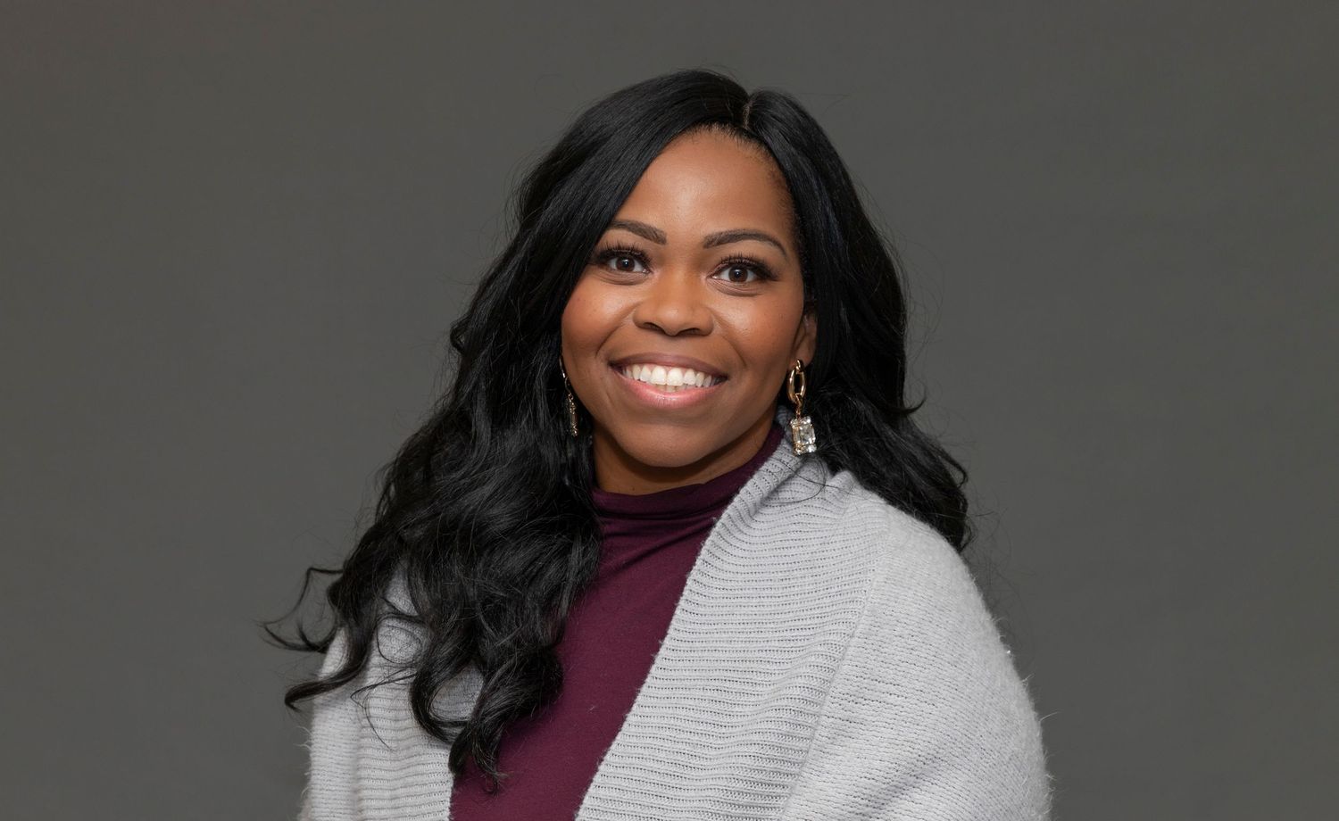 Professional headshot of a person wearing a burgundy top and grey cardigan against a dark background.