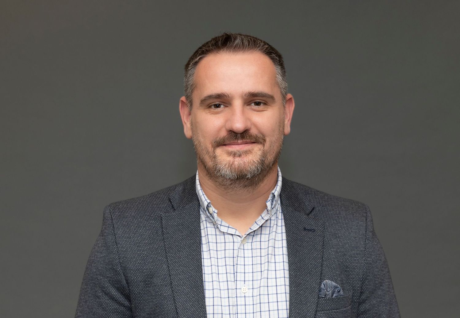 Professional headshot of a person in a grey suit jacket and checkered shirt against a dark background.