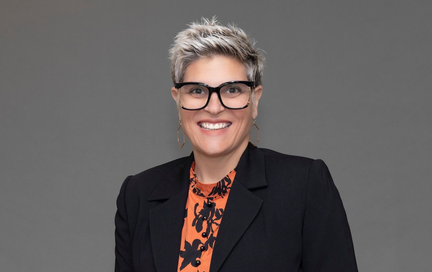 Professional headshot of a person with short grey hair wearing glasses and a patterned shirt.