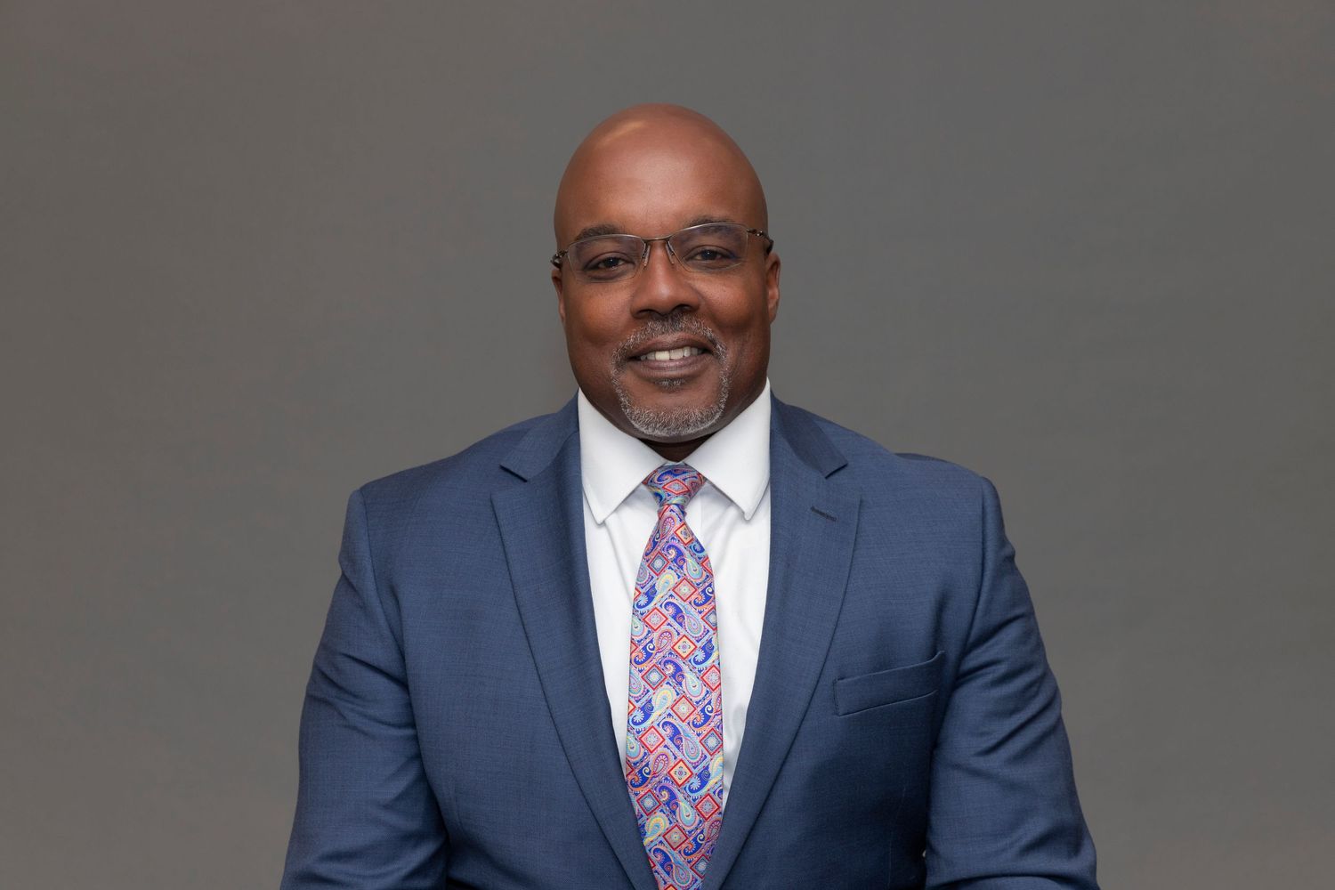 Professional headshot of someone wearing a navy suit with colorful floral tie against gray background.