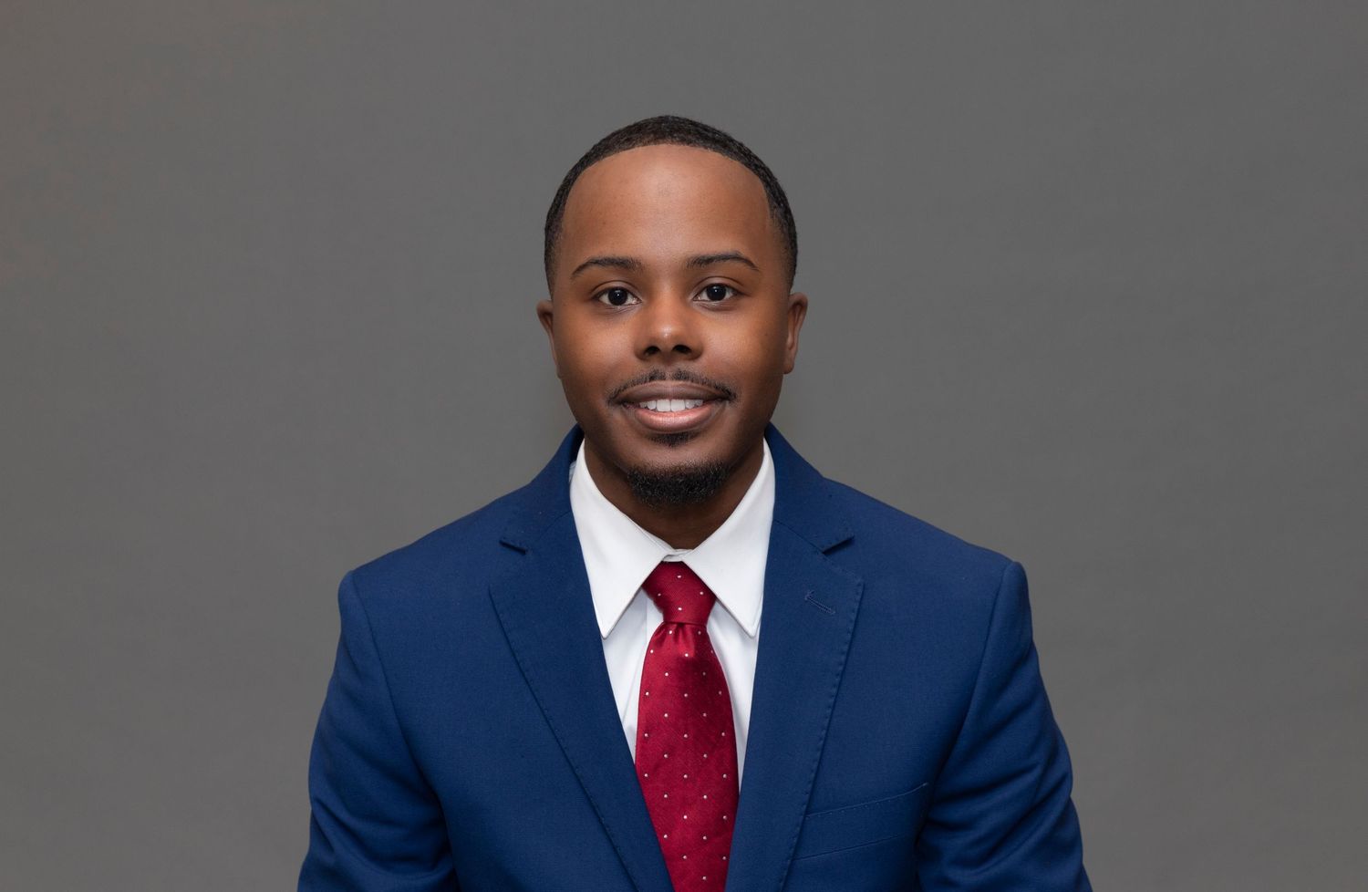 Professional portrait of person in navy suit and red tie against gray backdrop.