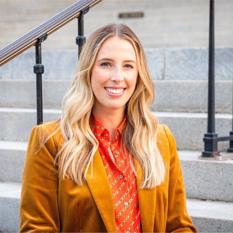 Professional portrait showing a bright smile, blonde wavy hair, orange blazer and patterned red blouse by steps.