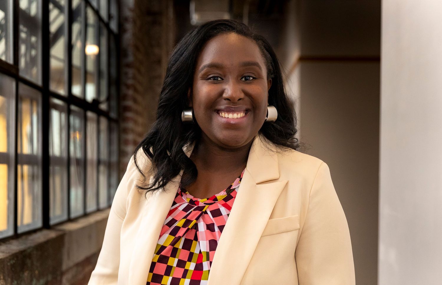Professional wearing a cream blazer and colorful patterned blouse smiles warmly in an office setting.