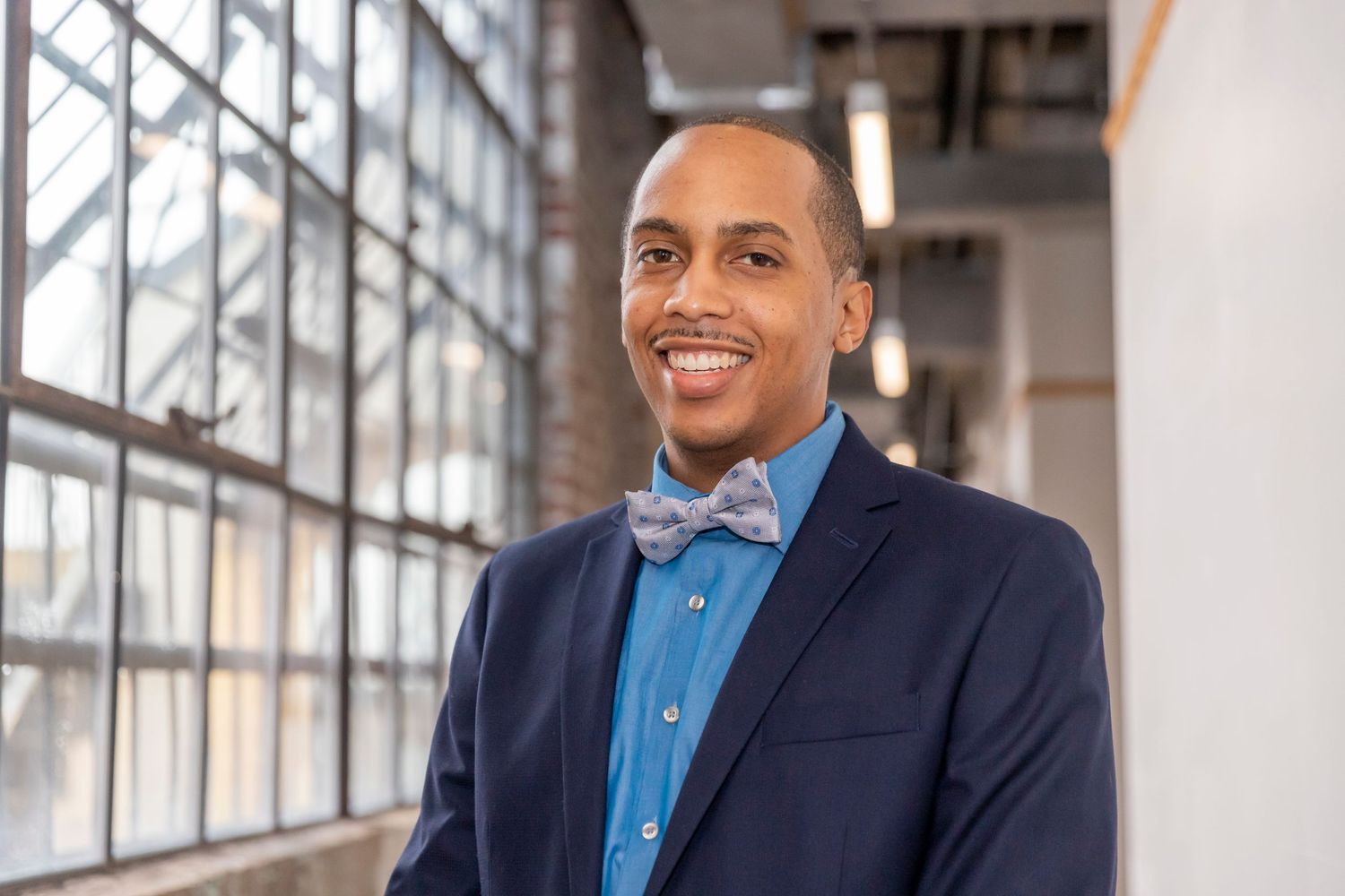 Professional in a navy blazer and light blue button-up shirt with bow tie stands near large windows in an office setting.