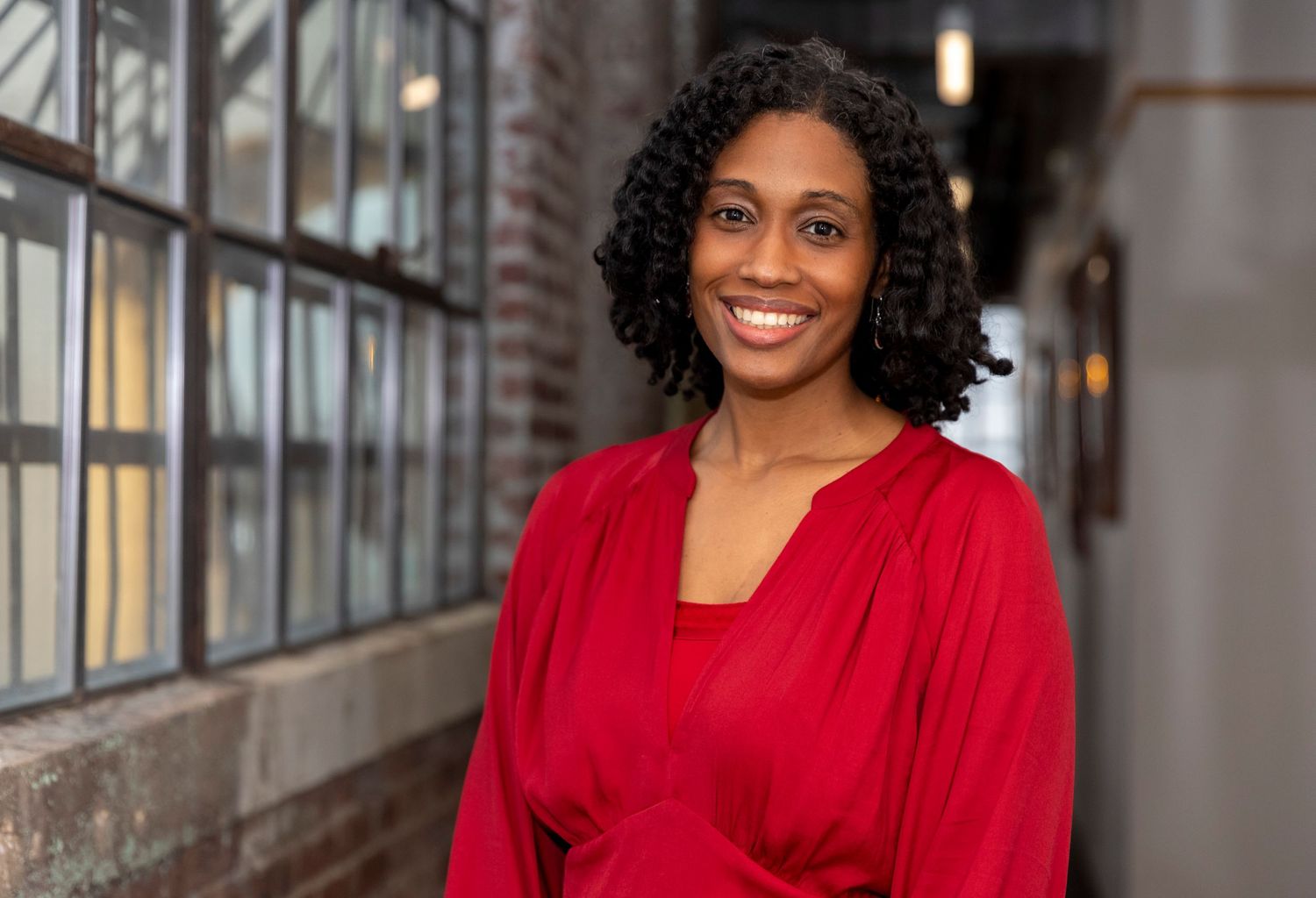 Professional business portrait of someone in a bright red blouse standing against a brick wall with windows.