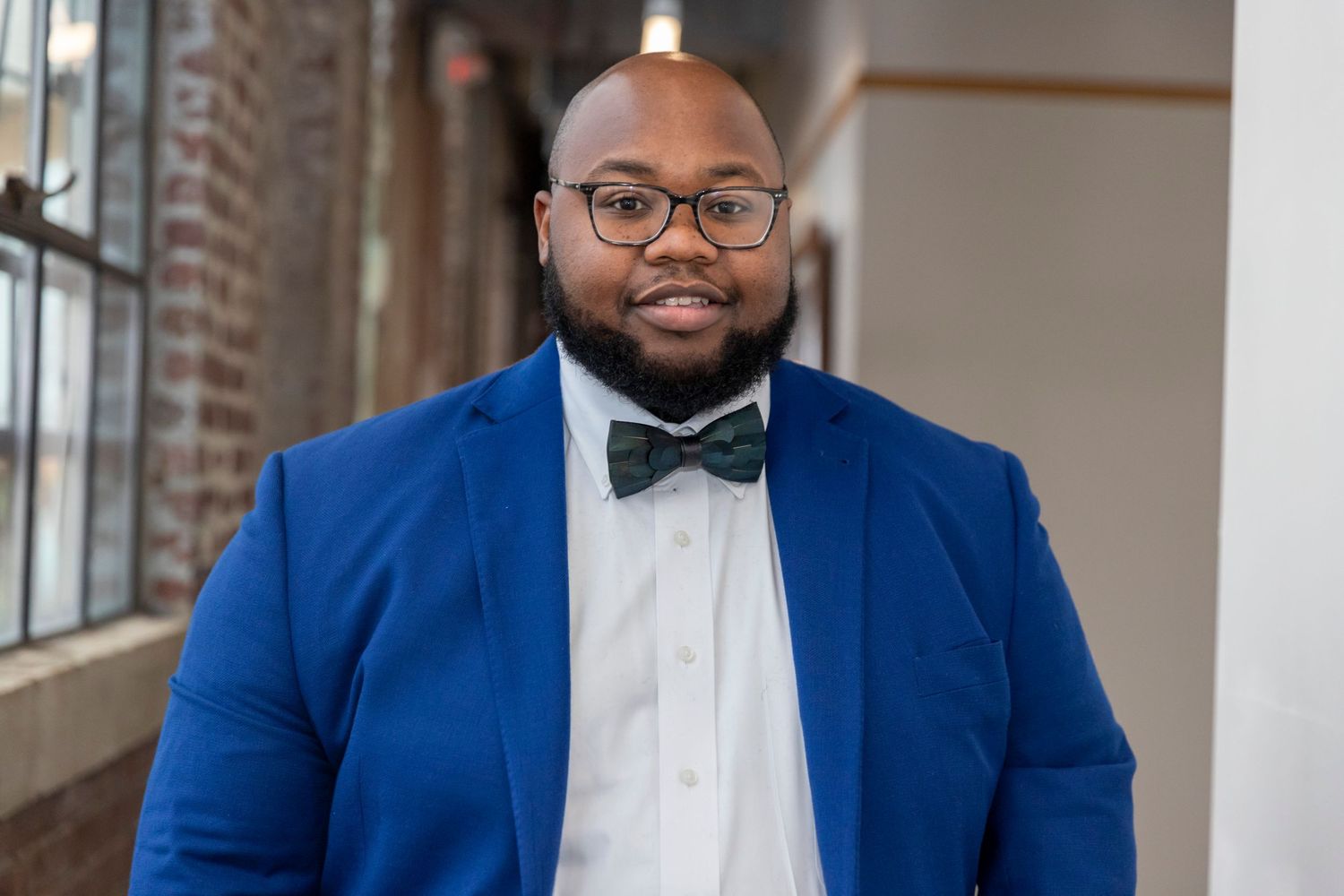 A professional in a royal blue suit jacket and bow tie smiles warmly while standing in a bright indoor setting.