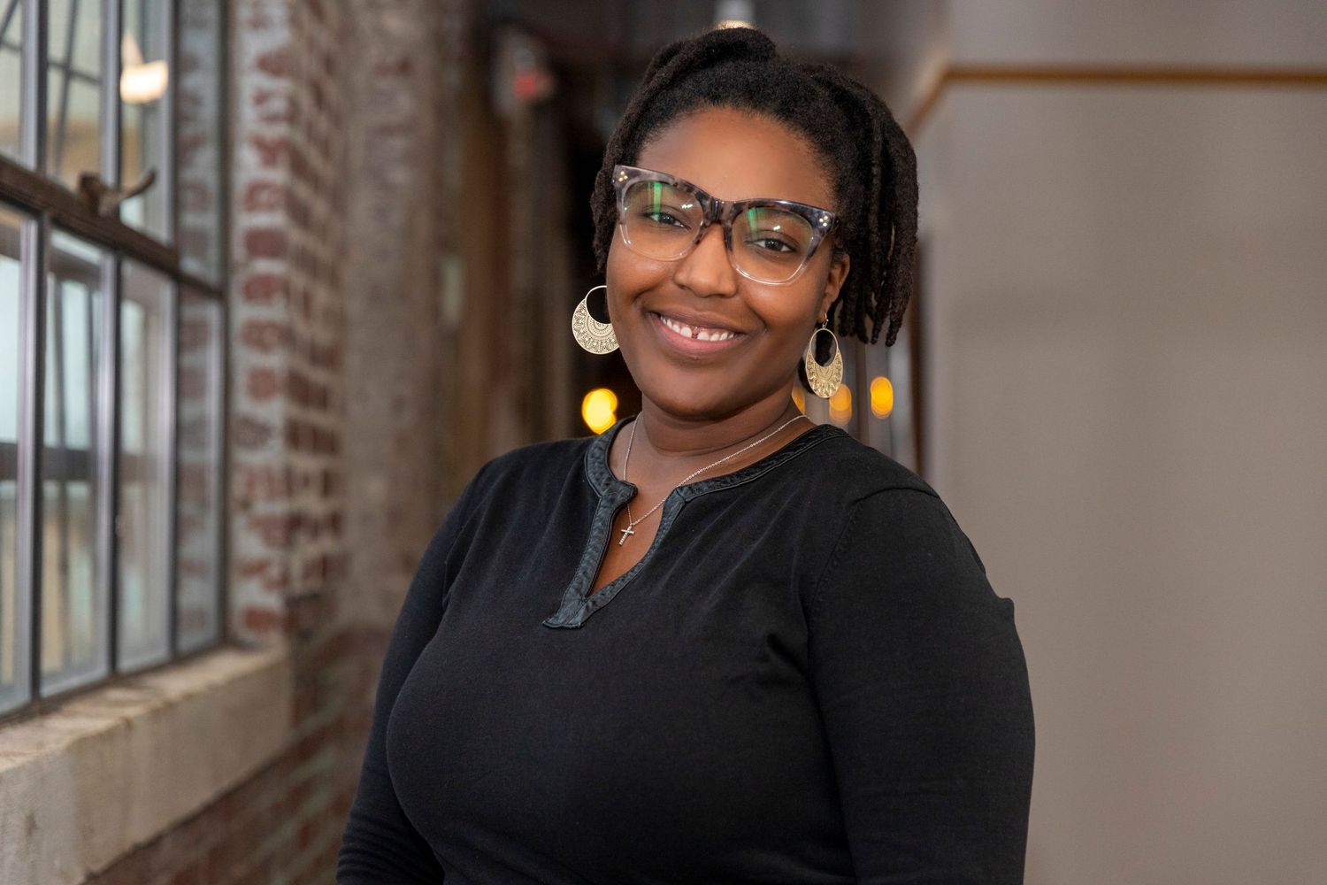 Professional headshot of someone wearing glasses and a black sweater smiling warmly against a blurred indoor background.