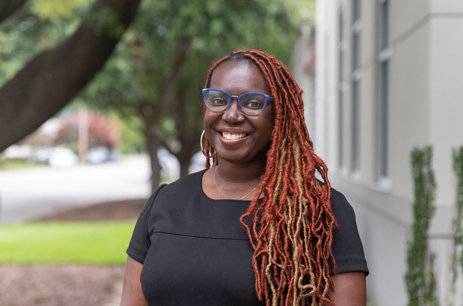 Portrait of someone with vibrant red locs wearing glasses and black top outdoors.