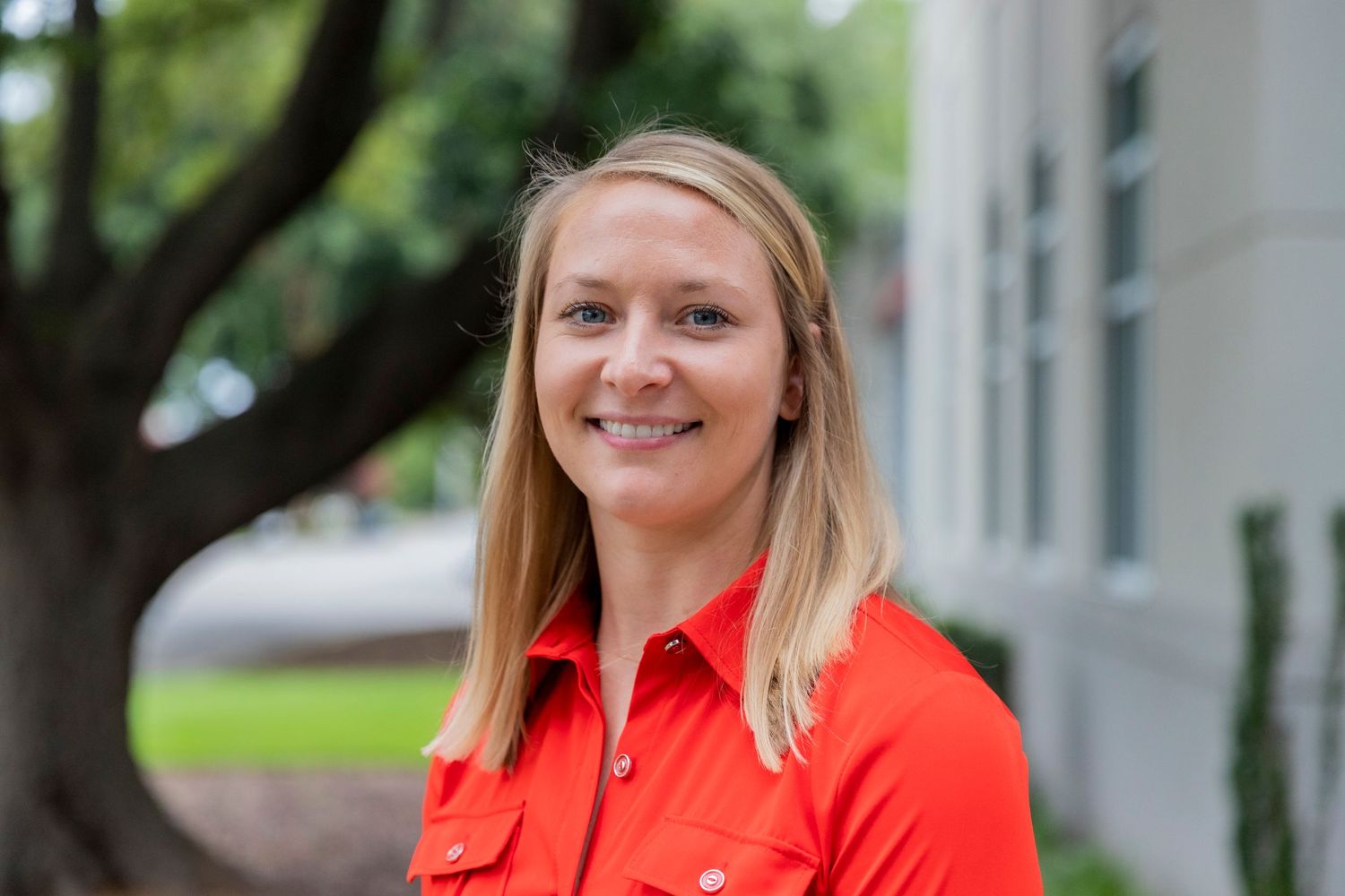 Professional headshot showing someone in a bright orange collared shirt outdoors against a blurred building exterior.